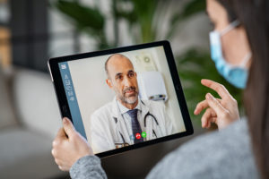 Sick young woman wearing protective mask during video call with mature doctor. Back view of patient with surgical face mask talking during conference call with her physician while staying at home in quarantine. Girl in online consultation with general practitioner on digital tablet.